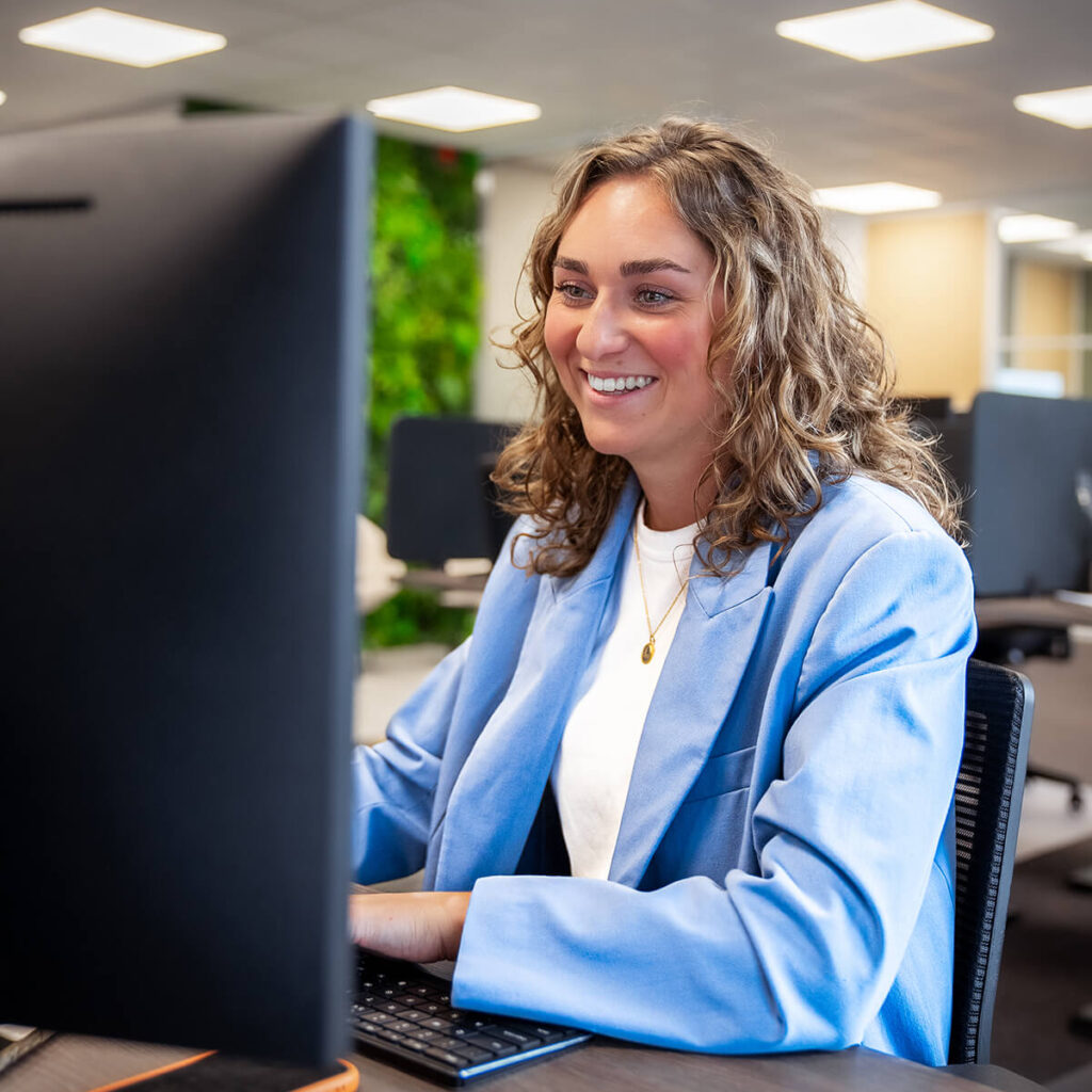 Vrouw in blauw jasje werkt glimlachend achter computer in modern kantoor