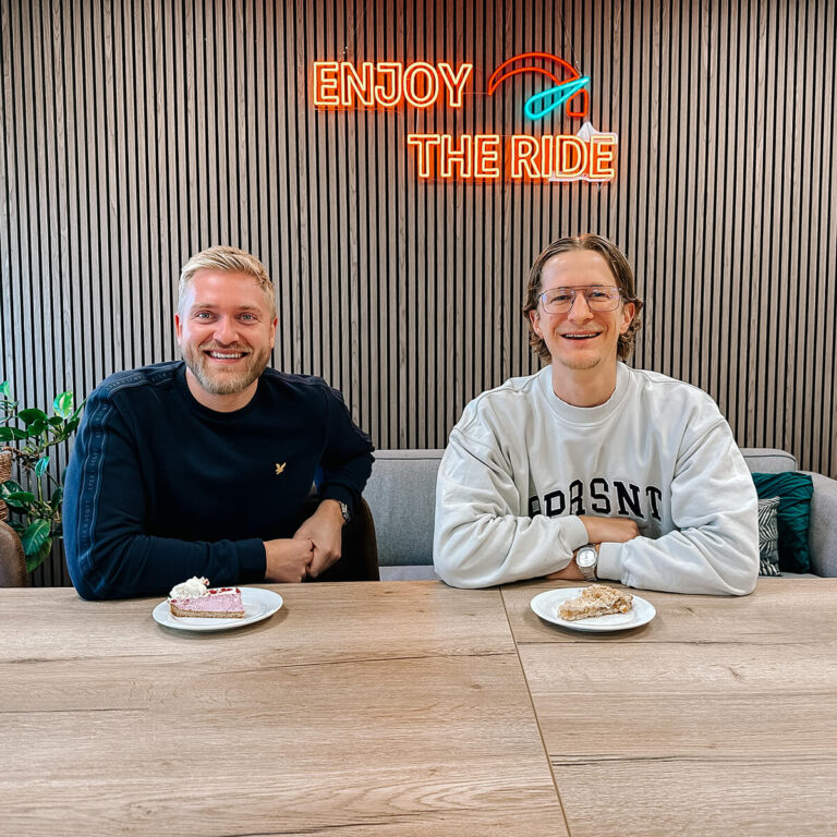 Twee lachende mannen zitten samen aan tafel met gebak in een modern kantoor